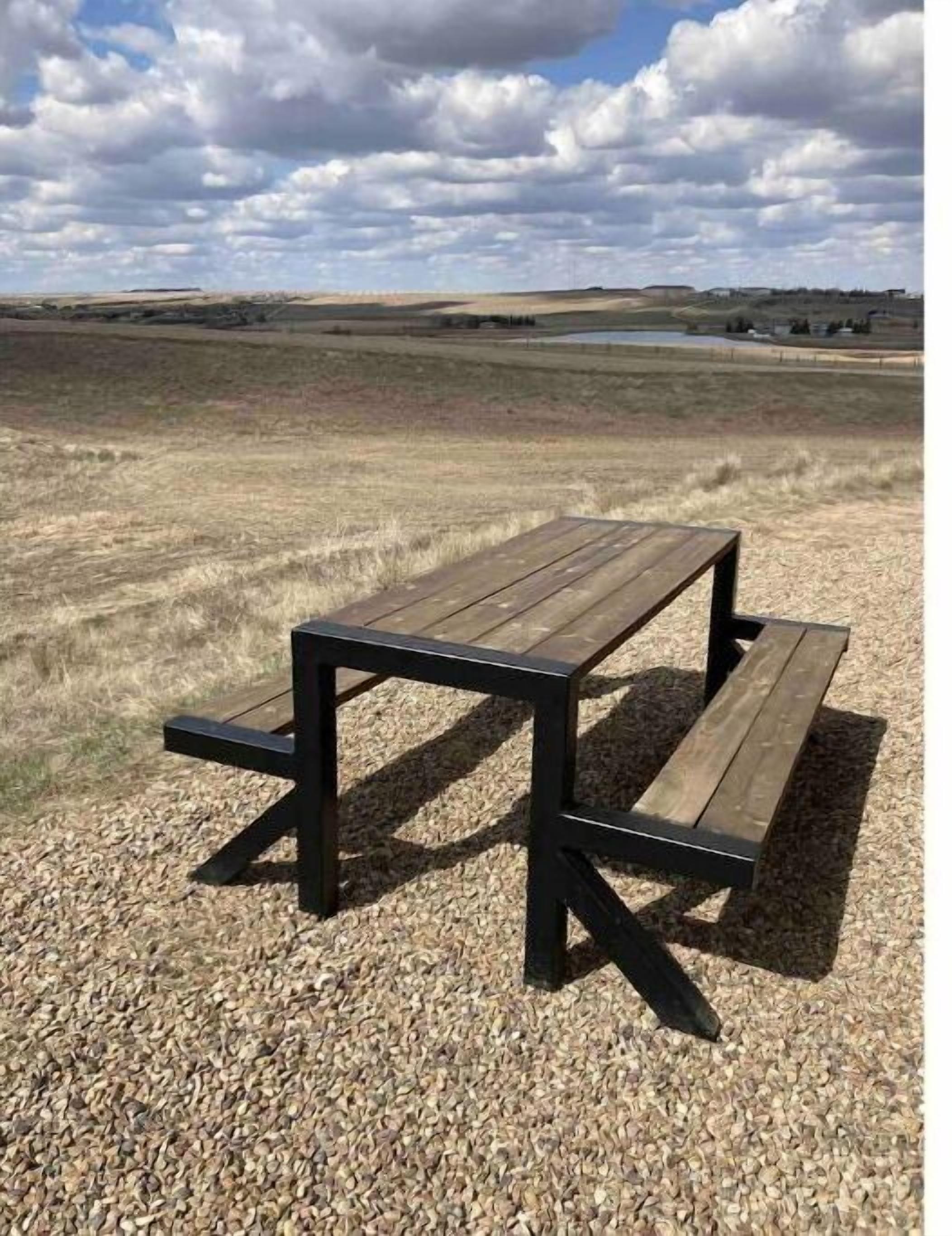 Une table de pique-nique en bois, avec des bancs, située dans un champ de gravier, d’où l’on aperçoit une étendue d’eau et des collines au loin.
