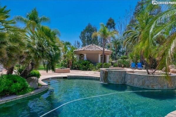 Relax by the serene pool surrounded by lush greenery and palm trees, featuring a gazebo and comfortable lounge chairs.