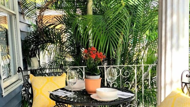Eine kleine Terrasse im Freien mit einem runden Tisch, zwei gelben Sitzgelegenheiten mit Kissen und einem Blumentopf mit roten Blüten.