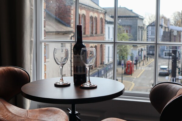 A table with a bottle and two glasses, a window with a view of buildings and a red telephone booth outside.