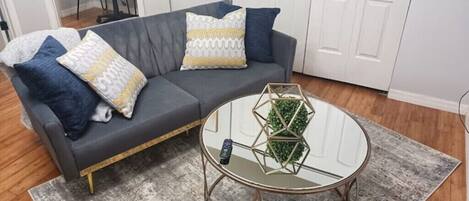 Un sofá gris con cojines estampados, una mesa de centro de cristal con una planta geométrica y un control remoto sobre la mesa.