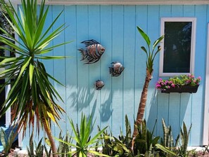 A blue house with three decorative fish mounted on the wall, a window with a flower box, and various tropical plants.