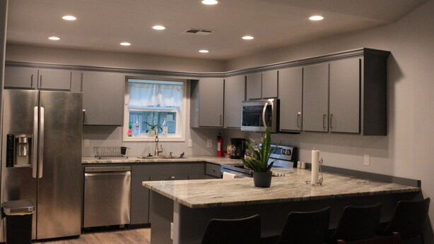 A modern kitchen with grey cabinets, stainless steel appliances, a central island with bar seating, and recessed ceiling lighting. - Chill Suburban Get Away (Corpus Christi)