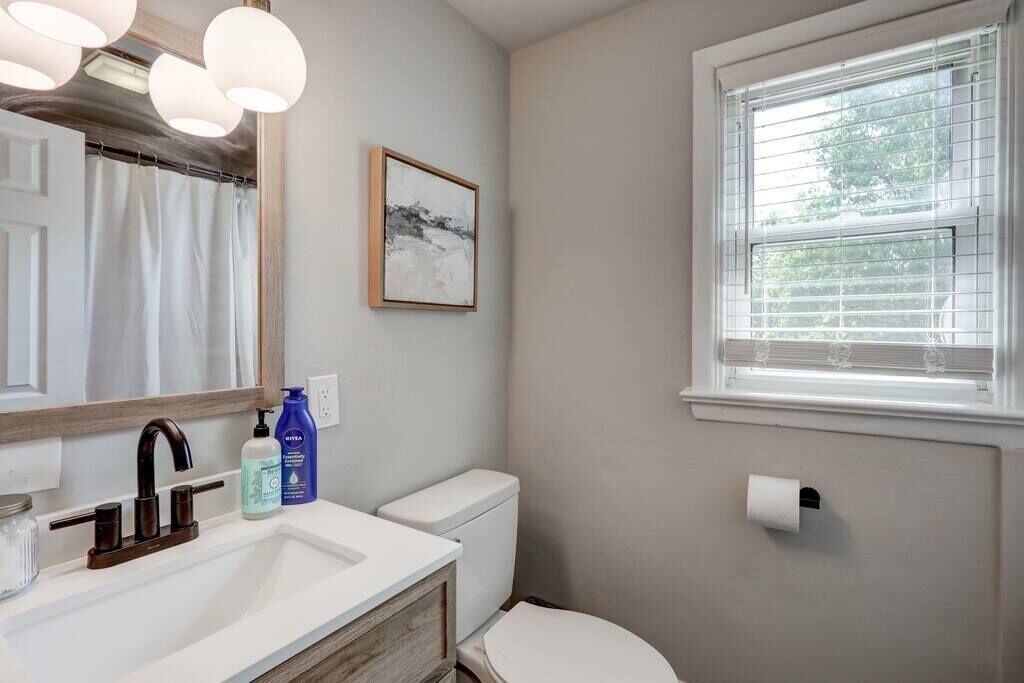 Une salle de bain avec un lavabo blanc, un robinet foncé et des toilettes. Il y a une fenêtre avec des stores, un tableau encadré au mur et un rouleau de papier toilette accroché au mur.