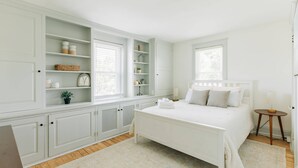 Une chambre avec un lit blanc, une armoire intégrée et une fenêtre avec des stores.