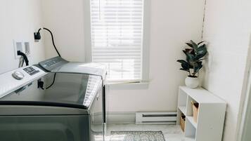 A laundry room with a washing machine, a window with blinds, a potted plant, and a white shelf.