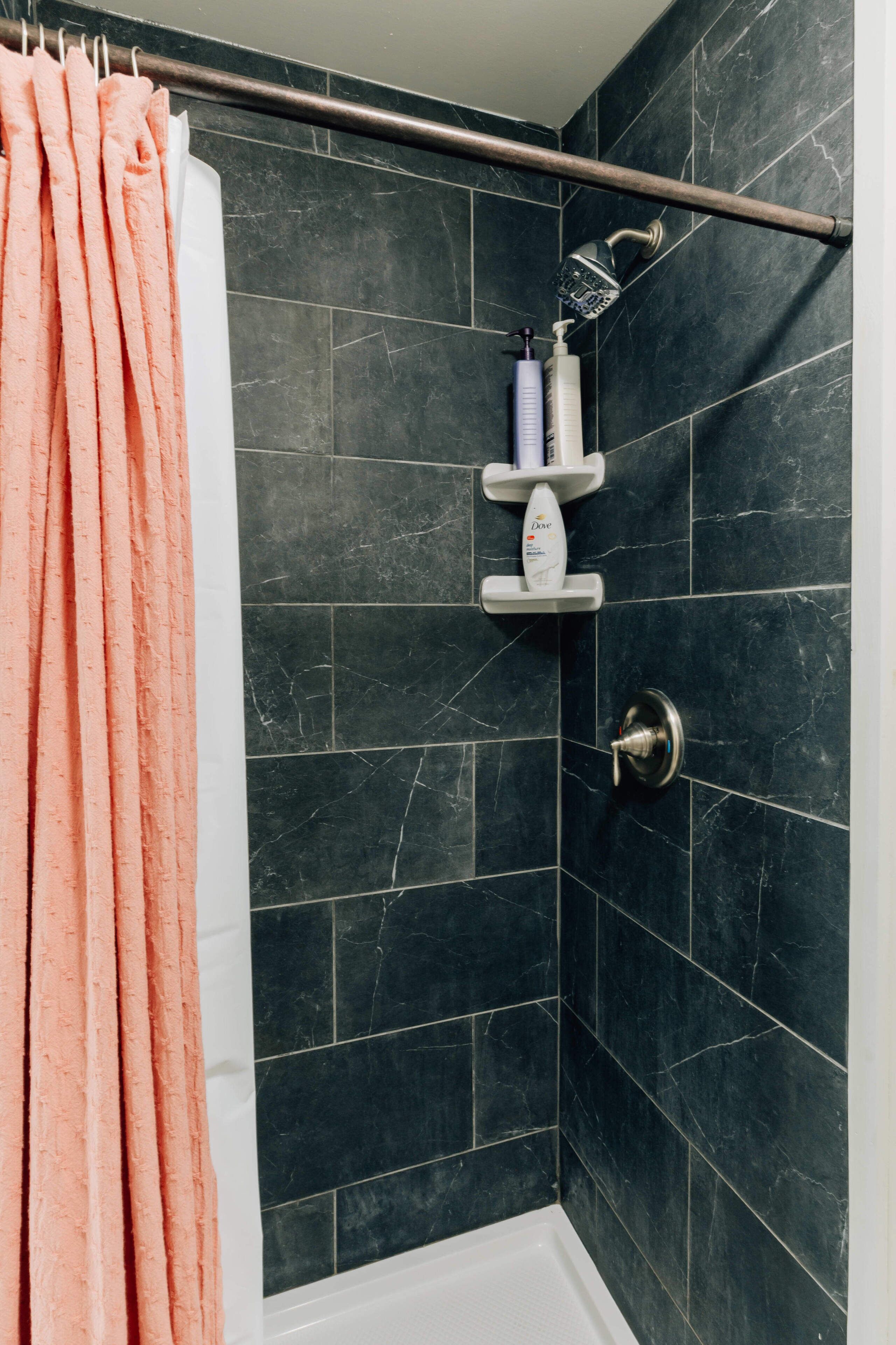 Ein Badezimmer mit dunkel gefliesten Wänden, einer weißen Badewanne, einer Dusche mit Duschbrause und einem Handtuchhalter mit einem rosafarbenen Handtuch.
