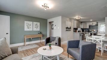 A modern living room with a sofa, armchair, and coffee table, a dining area with a table and chairs, and a kitchen with cabinets and appliances.
