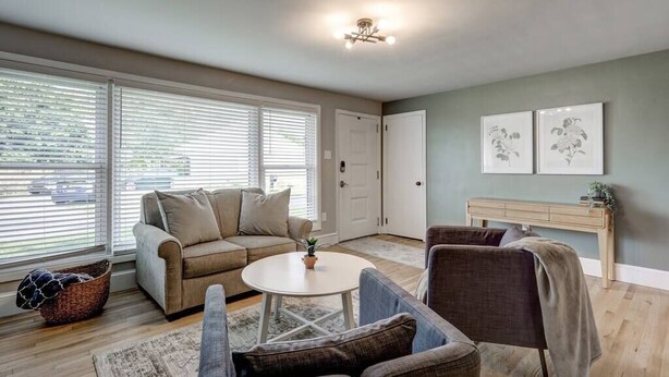 A living room with a beige sofa, a round coffee table, and a grey armchair. There are two framed pictures on the wall, a white door, and a window with blinds. - New cozy home near Outlets & Amish Country (Lancaster)