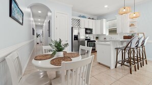 A modern kitchen with white cabinets, a central island, and a dining area with white chairs and woven placemats. - Newly Remodeled 2BR/2B Condo (South Padre Island)