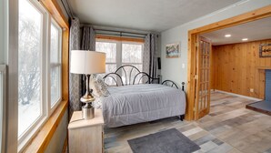 A bedroom with a bed, a bedside table with a lamp, a window with curtains, and a wooden door leading to another room.