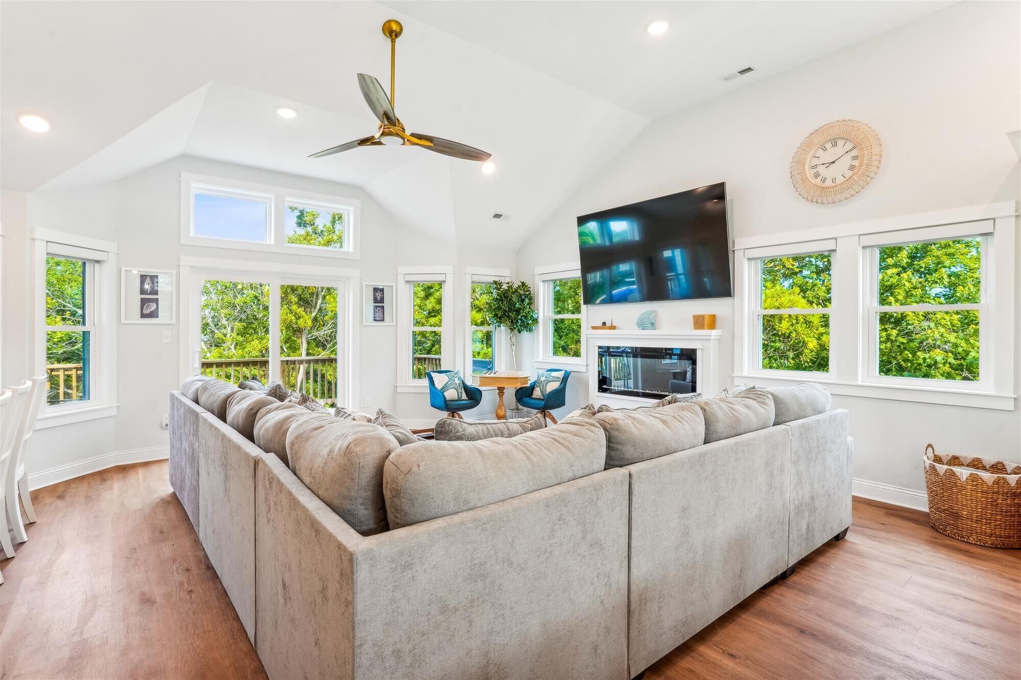 A spacious living room with a large sectional sofa, a ceiling fan, a flat-screen TV, and large windows offering a view of greenery.