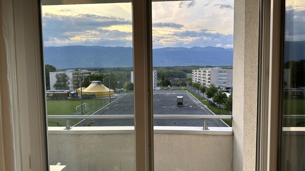 A view from a window overlooking a parking lot, a yellow tent, and buildings in the distance with mountains in the background. - Quartier de Meyrin (Meyrin)