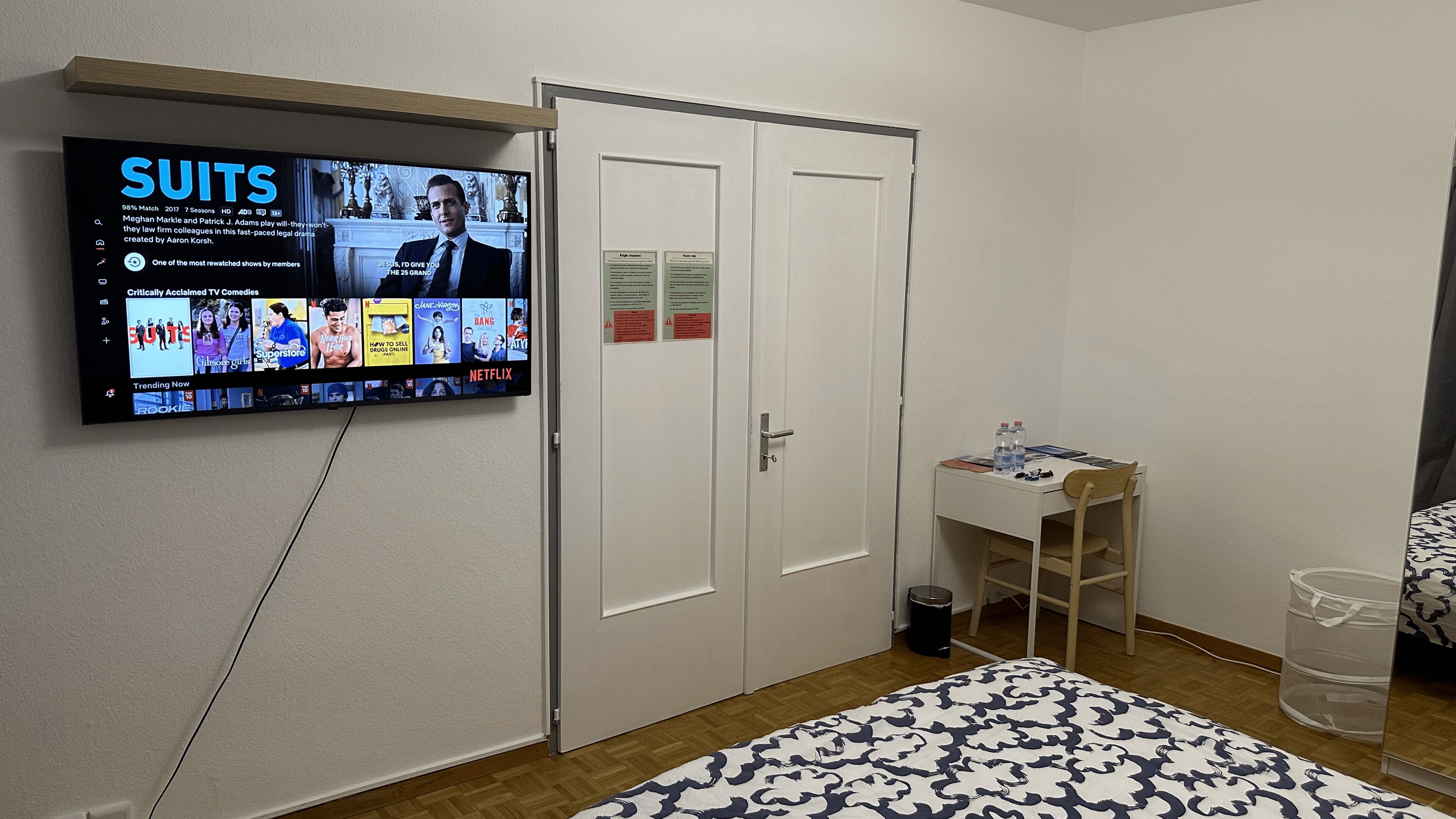 A hotel room with a TV mounted on the wall displaying 'SUITS' and other shows, a white door, a small desk with a chair, and a patterned bedspread.