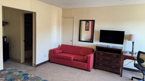 A hotel room with a bed, a red sofa, a TV, a desk with a lamp, and a dresser. - Village Inn (Ada)