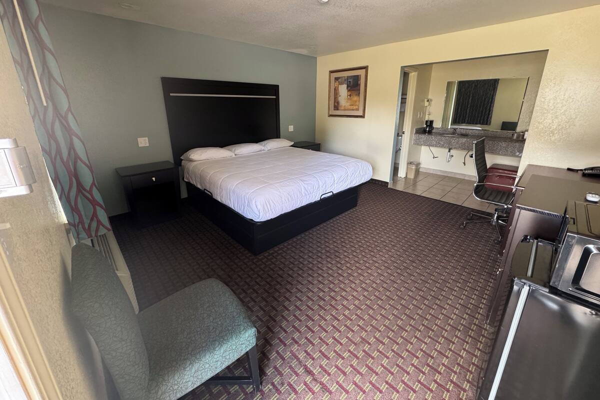 A hotel room with a bed, a desk, a chair, a bench, a bathroom, and a mirror.
