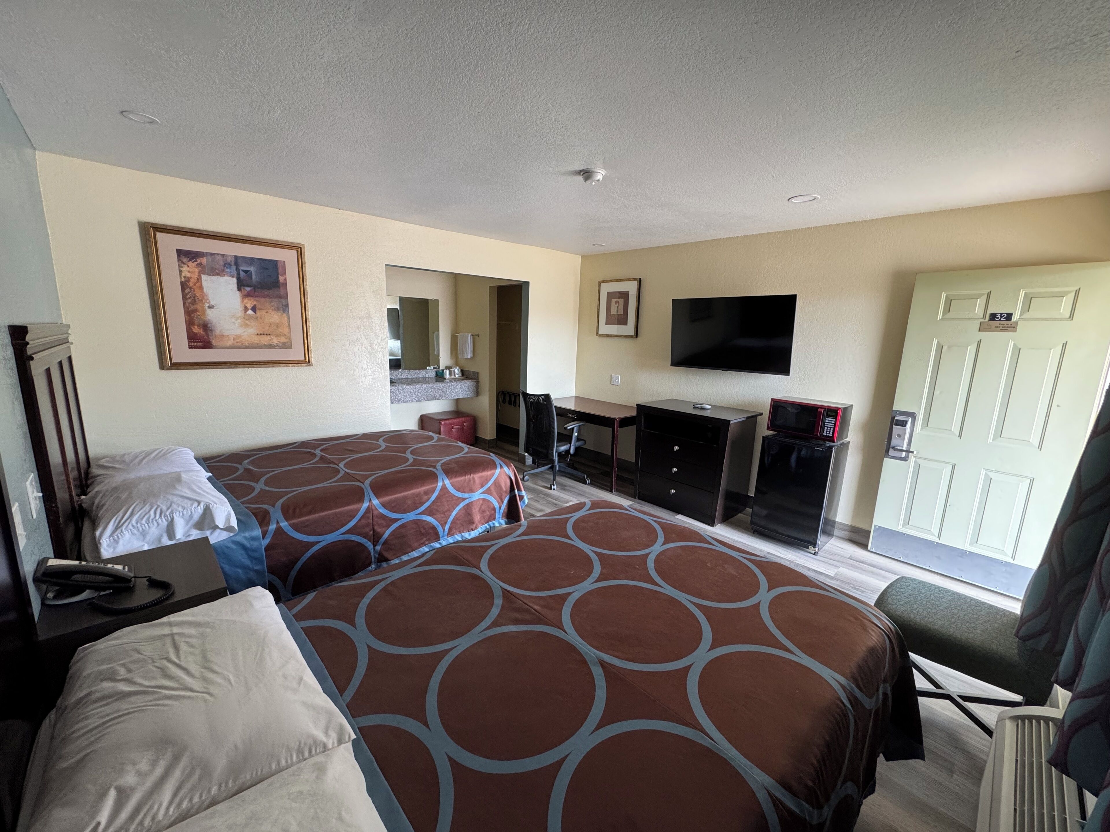 A hotel room with two beds, a desk, a chair, a television, a microwave, and a door with a number.