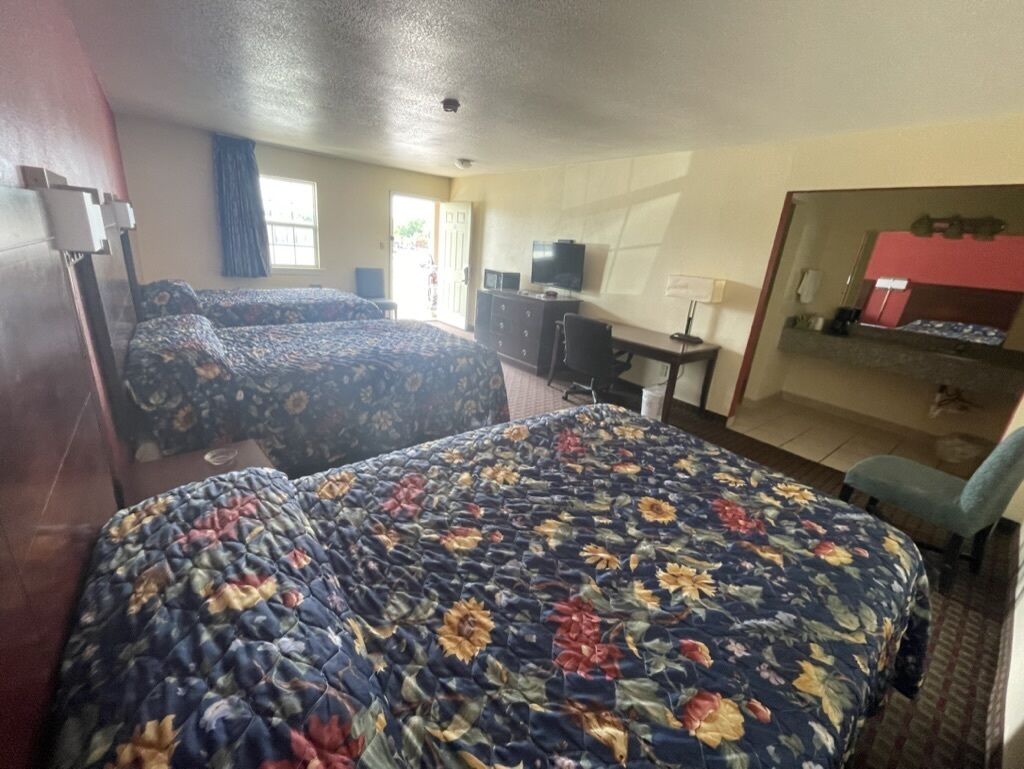 A hotel room with two beds, a desk, a chair, a television, and a bathroom visible through an open door.