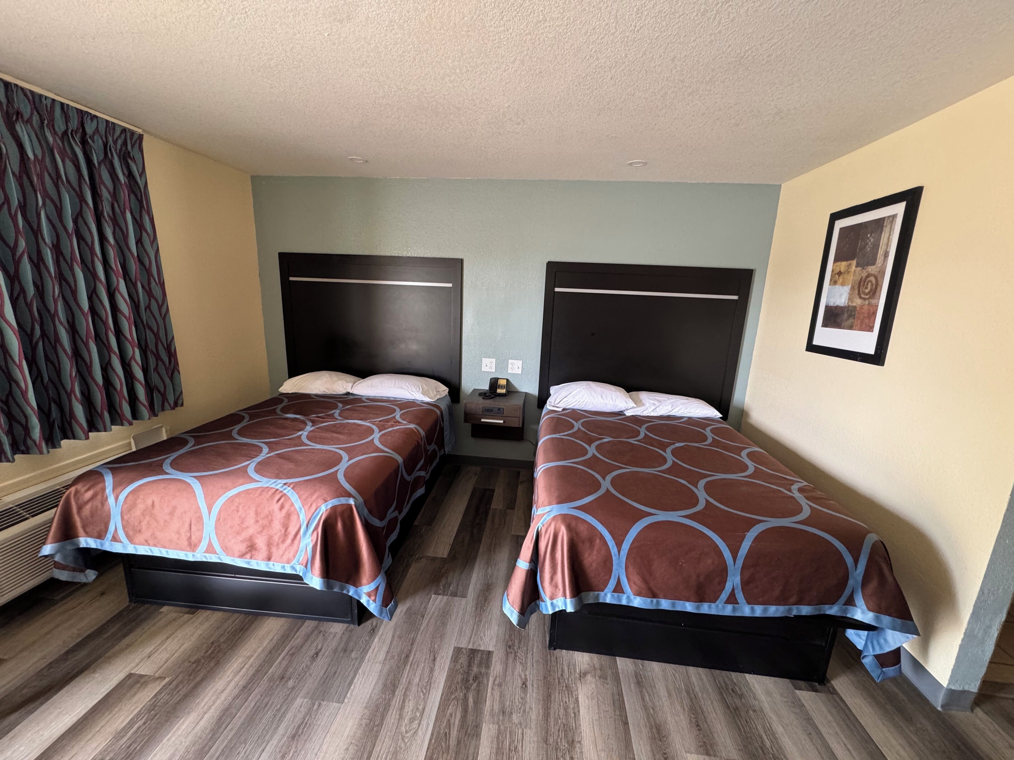 A hotel room with two beds, a nightstand, a wall-mounted phone, and a framed picture.