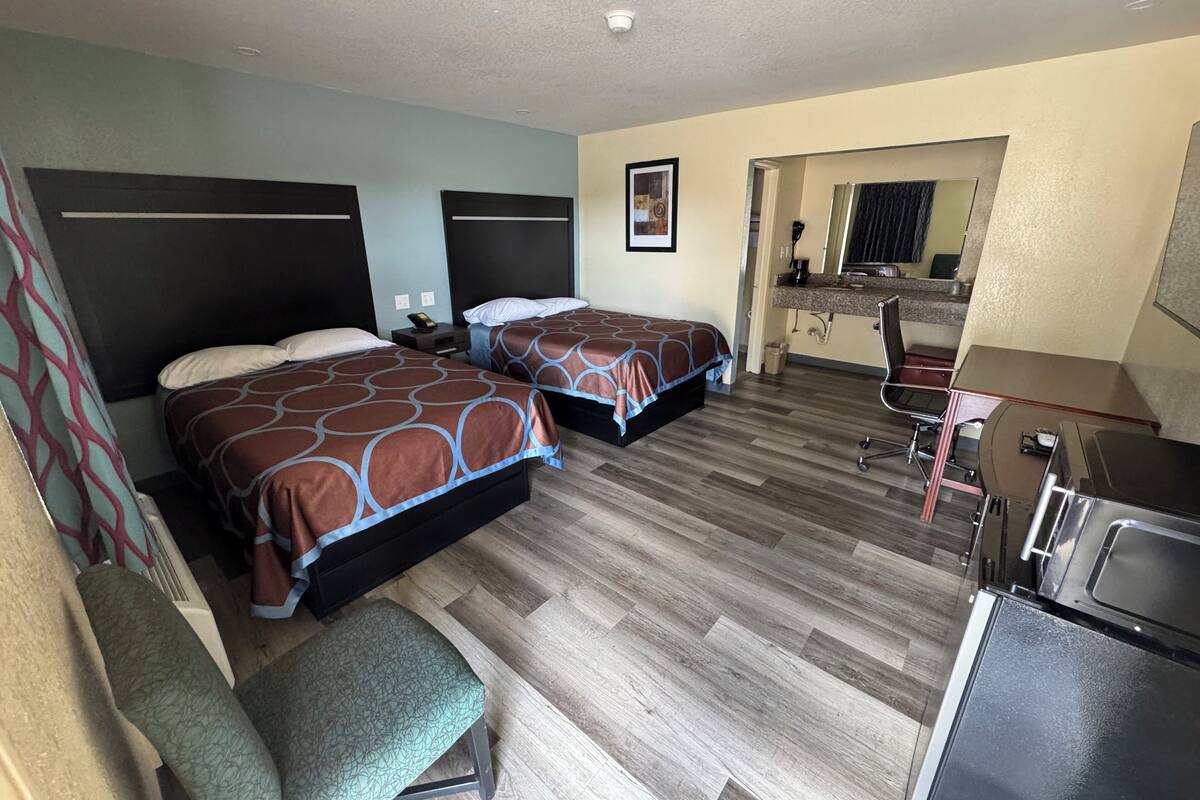 A hotel room with two beds, a desk, a chair, a kitchenette, and a bathroom visible through an open doorway.