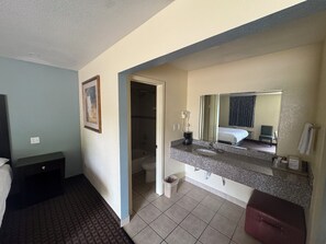 A hotel room with a bed, a desk, a bathroom with a sink and mirror, and a shower.