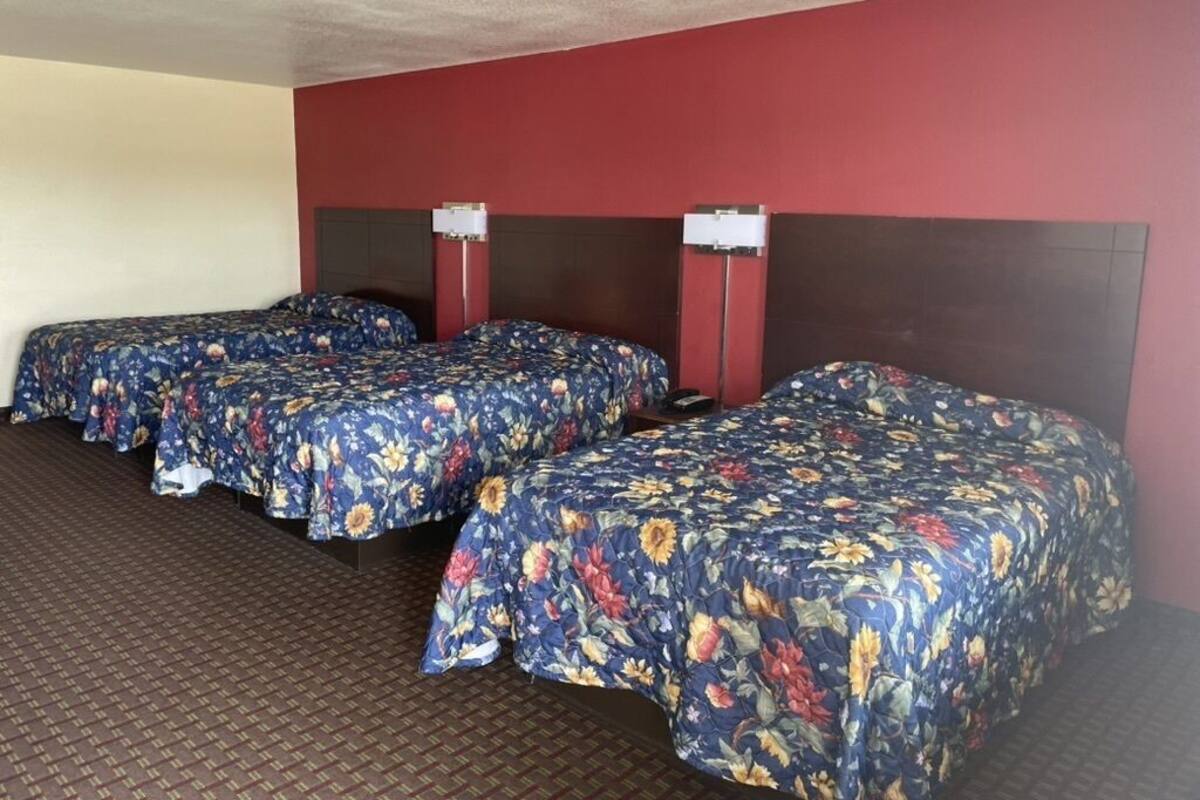 A hotel room with three single beds, each with a floral bedspread, a dark headboard, and a nightstand with a lamp.