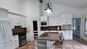 A spacious kitchen with white cabinetry, a central island, and a large window providing natural light. - Ranch House (Alba)