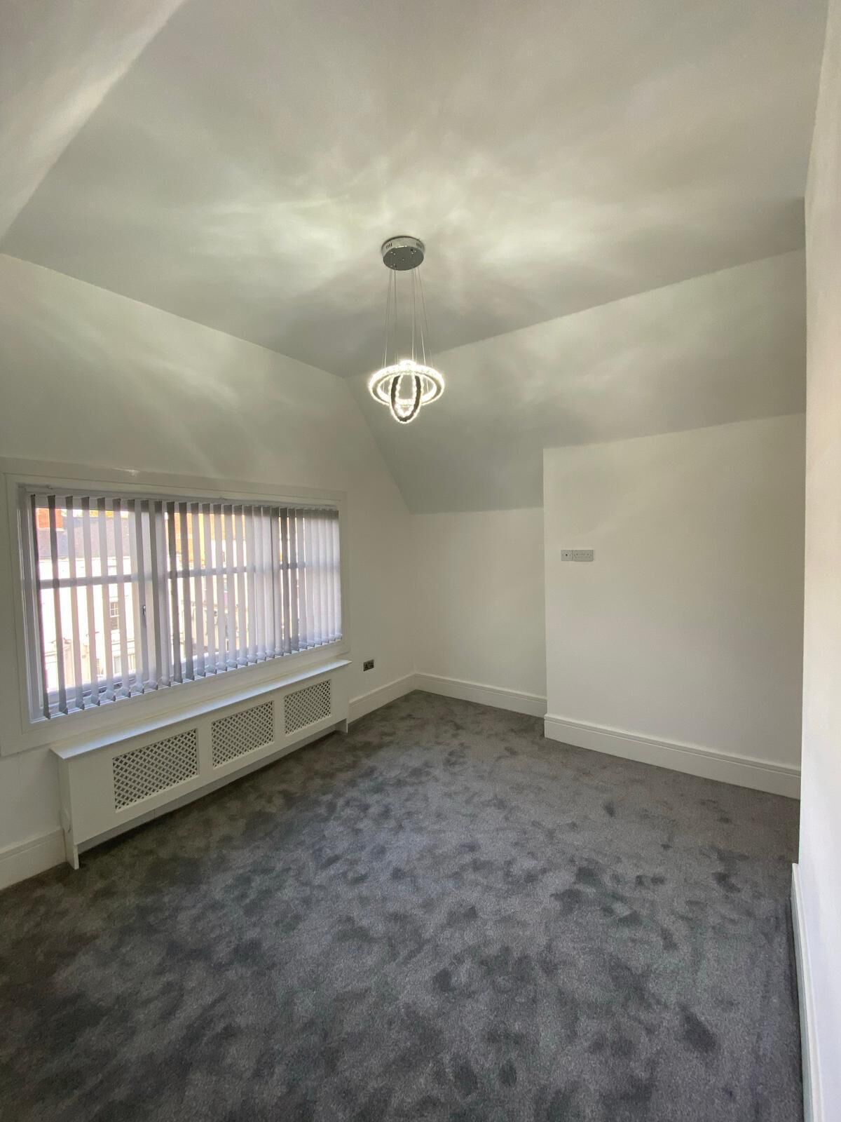 A room with a sloped ceiling, a window with blinds, a radiator, and a carpeted floor.