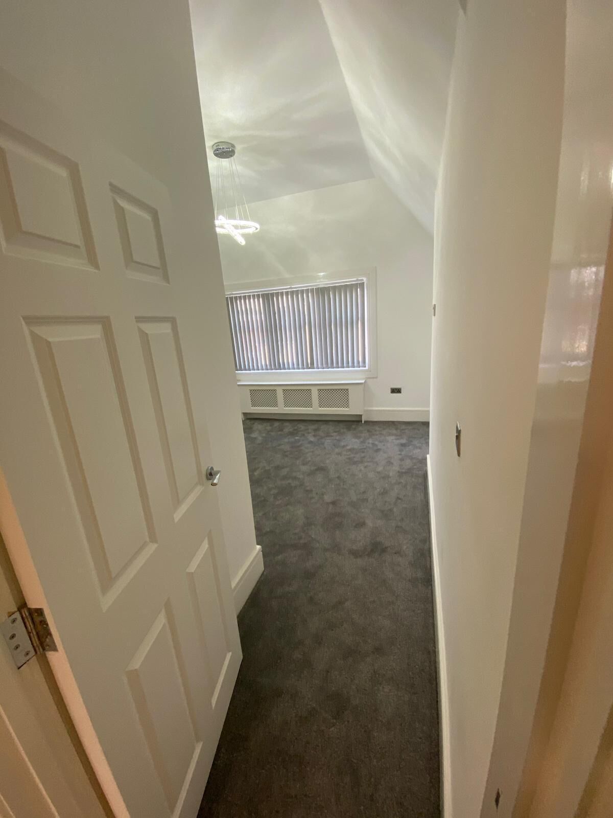 A narrow hallway with a carpeted floor, white walls, and a window with blinds.