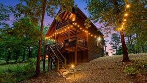 A wooden cabin with a balcony, surrounded by trees and gravel path. - Forward My Calls · Elevate Your Stay: 4 Decks, Hot Tub, & Idyllic Outdoor Haven (Broken Bow)