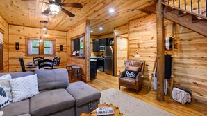 A wooden cabin interior with a sofa, a chair with a 'Hello' pillow, a dining table, and a kitchen area. - Forward My Calls · Elevate Your Stay: 4 Decks, Hot Tub, & Idyllic Outdoor Haven (Broken Bow)