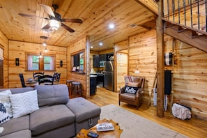 A wooden cabin interior with a sofa, a chair with a 'Hello' pillow, a dining table, and a kitchen area.