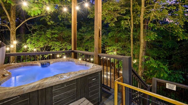 A hot tub in a wooden deck area surrounded by trees, with string lights overhead. - Forward My Calls · Elevate Your Stay: 4 Decks, Hot Tub, & Idyllic Outdoor Haven (Broken Bow)