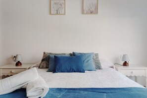 Un lit bien fait, avec une couverture bleue et des oreillers, deux tables de chevet avec des lampes, et deux tableaux encadrés accrochés au mur.