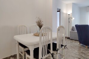 Un coin repas avec une table et des chaises blanches, un vase contenant des fleurs séchées et une lampe en arrière-plan.