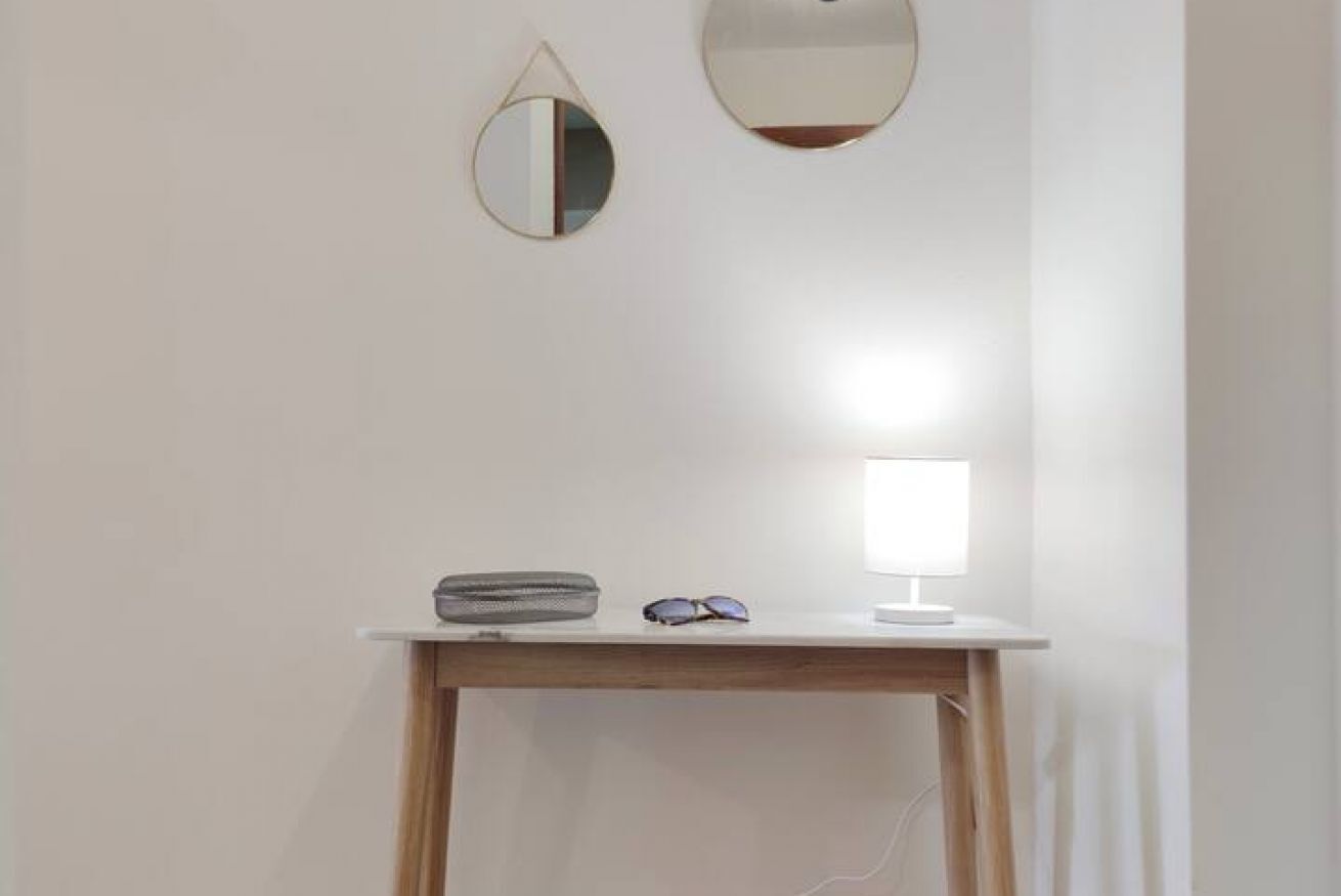 Ein Zimmer mit weißer Wand, einem kleinen Holztisch, einer Schreibtischlampe, einer Sonnenbrille und zwei Wandspiegeln.