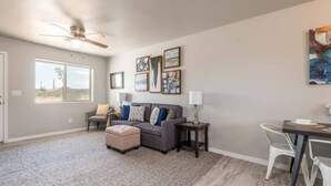 A living room with a gray sofa, a small ottoman, a dining table with chairs, and a ceiling fan. - Brand New Duplex! Minutes from every amenity! (Kingman)