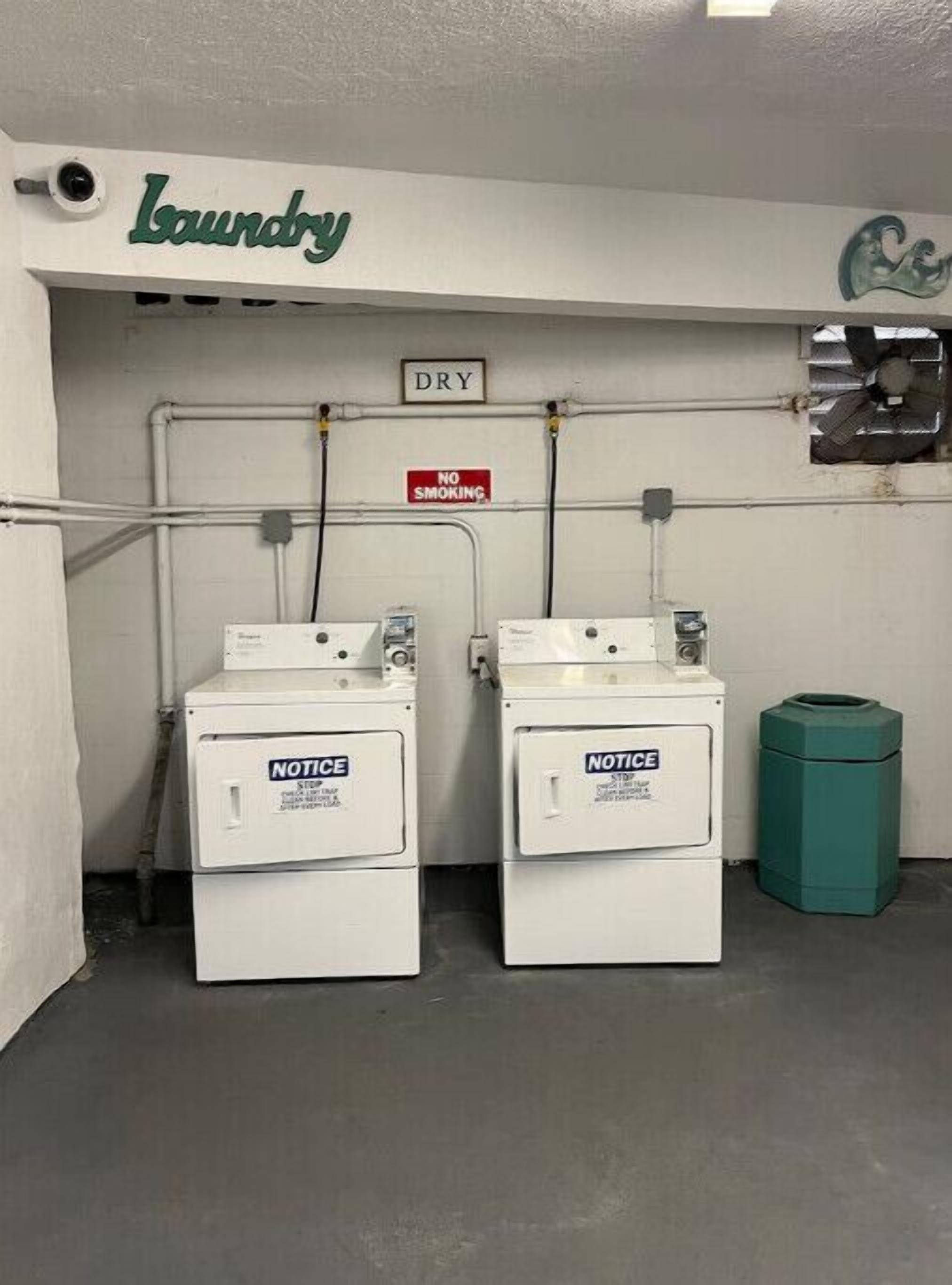 A laundry area with two washing machines, a 'No Smoking' sign, and a 'Dry' sign above a damaged window.