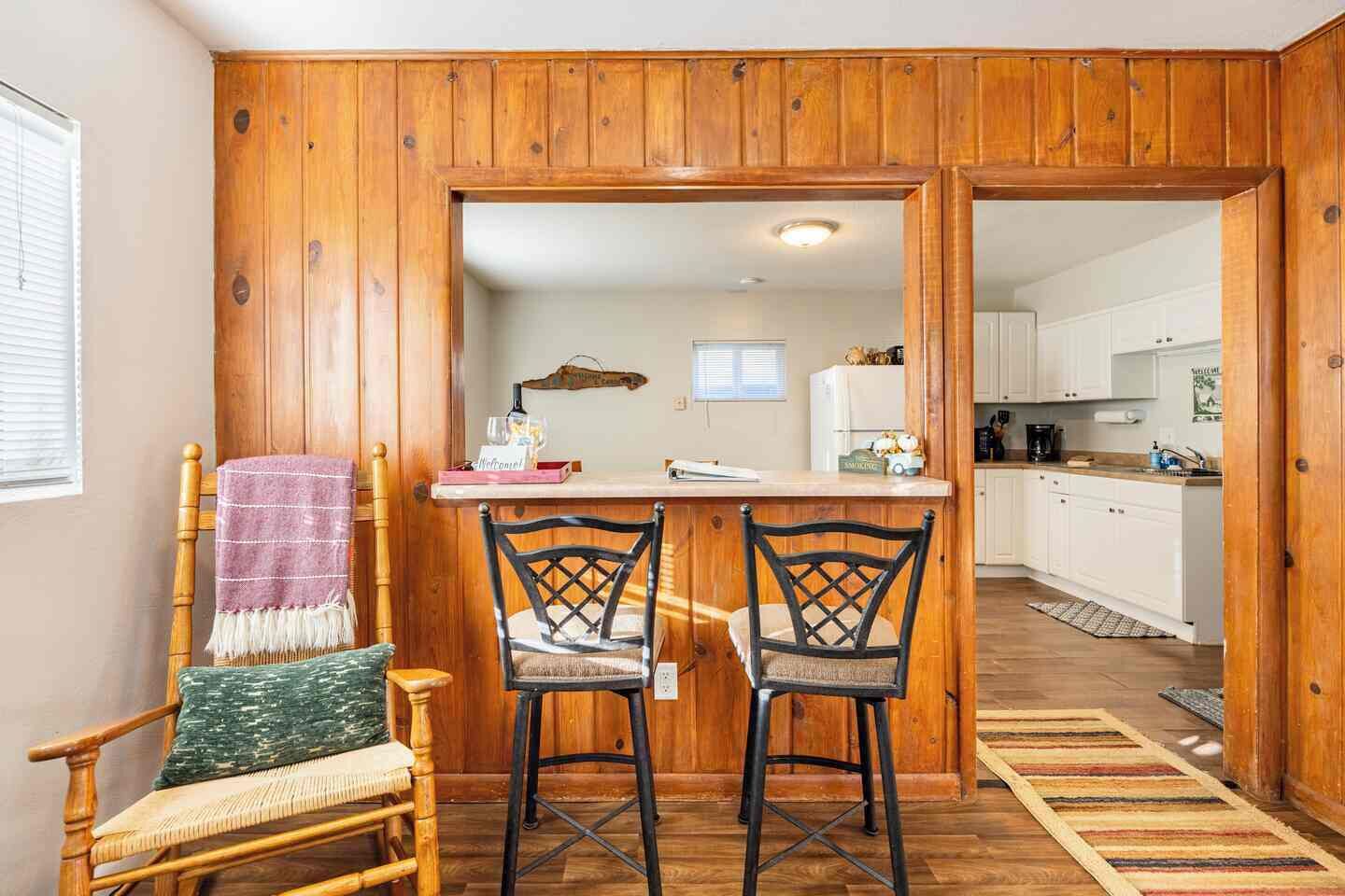 A wooden-paneled room with a kitchen area, two bar stools, a wooden chair, and a small table with a glass.