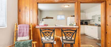 A wooden-paneled room with a kitchen area, two bar stools, a wooden chair, and a small table with a glass.