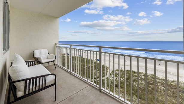 A balcony with a white railing, two white cushioned chairs, and a view of the beach and ocean.