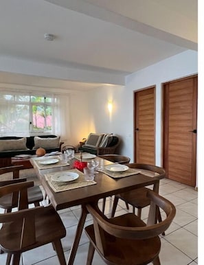 Un coin repas avec une table dressée pour quatre personnes, entourée de chaises en bois. On aperçoit un canapé et un téléviseur au fond.
