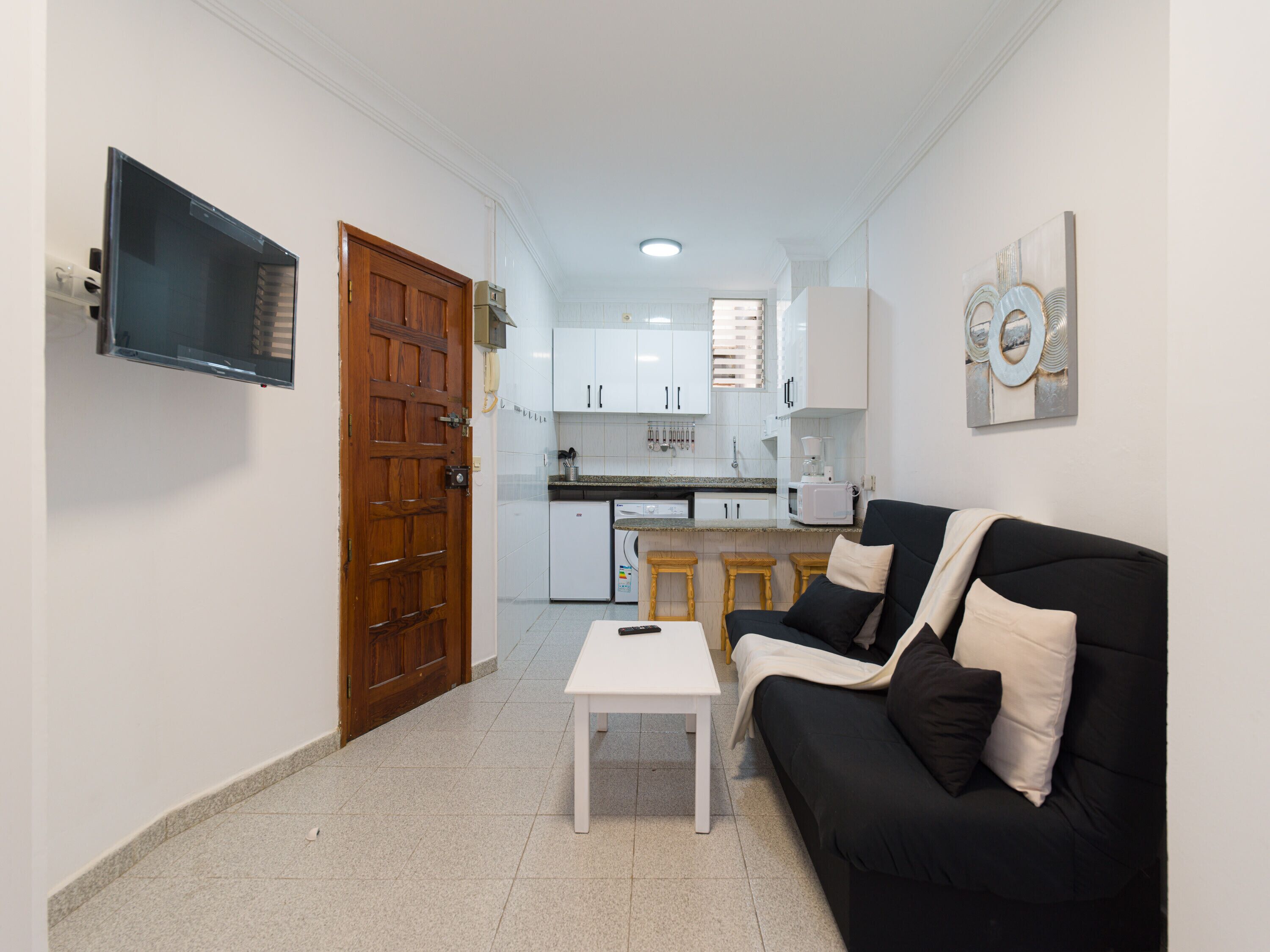 A compact living space with a kitchenette, a black sofa with white cushions, a small white table, and a TV mounted on the wall.