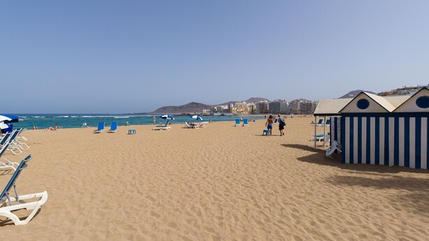 Ein Strand mit blauem Himmel, blauen Sonnenschirmen und Liegestühlen.