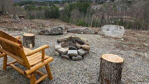 En træstol og et ildsted i sten i et naturligt landskab med udsigt til skov og bjerge.