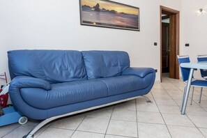 Um sofá azul em um quarto com piso de azulejos, um quadro na parede e uma porta que dá para outro cômodo.