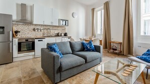 A modern kitchen with white cabinets, a built-in oven, and a microwave. There is a grey sofa with blue pillows in the living area, and a glass coffee table.