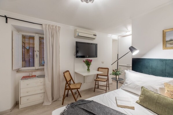 A bedroom with a bed, a desk with a chair, a TV, an air conditioner, and a window with curtains.