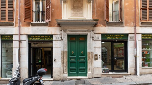 A building with a green door, storefronts labeled 'FERRAMENTA MATERIALE EDILE' and 'CASALINGHI DETERSIVI,' and a parked scooter. - GuestHost - Basic Studio - 350 m far from Fontana di Trevi (Roma)