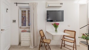 A small room with a white desk, two wicker chairs, a flat-screen TV, an air conditioning unit, and a window with curtains. - GuestHost - Basic Studio - 350 m far from Fontana di Trevi (Roma)